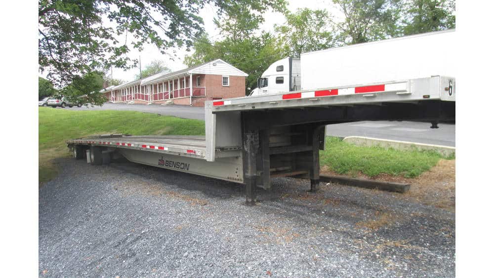 Flatbed trailer with black and silver color parked on gravel surface near a residential building.