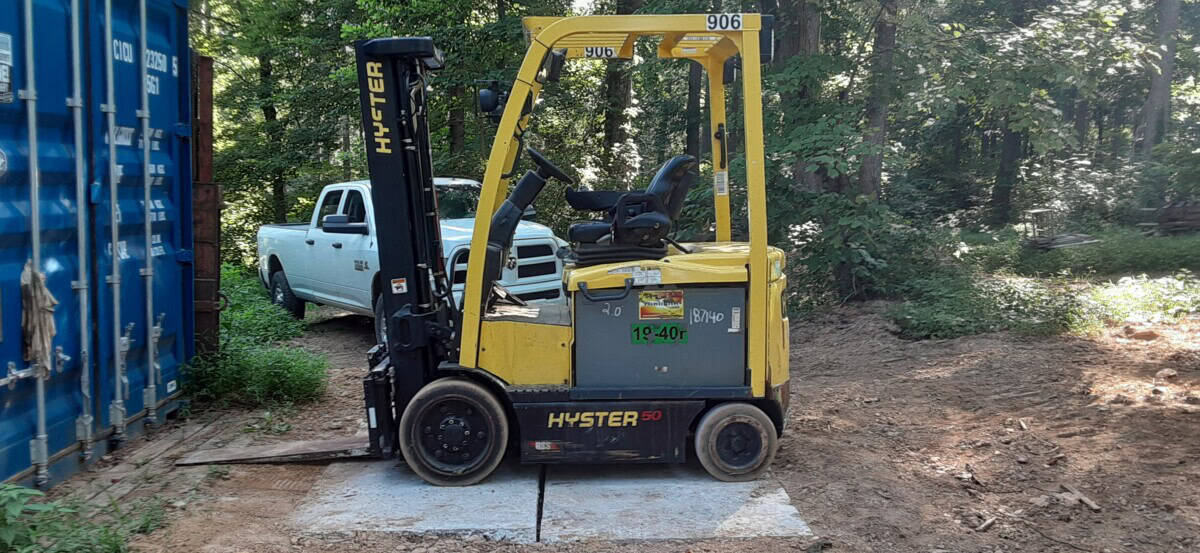 Yellow Hyster E50XM forklift with black mast and tires, parked on dirt near trees and a blue shipping container.