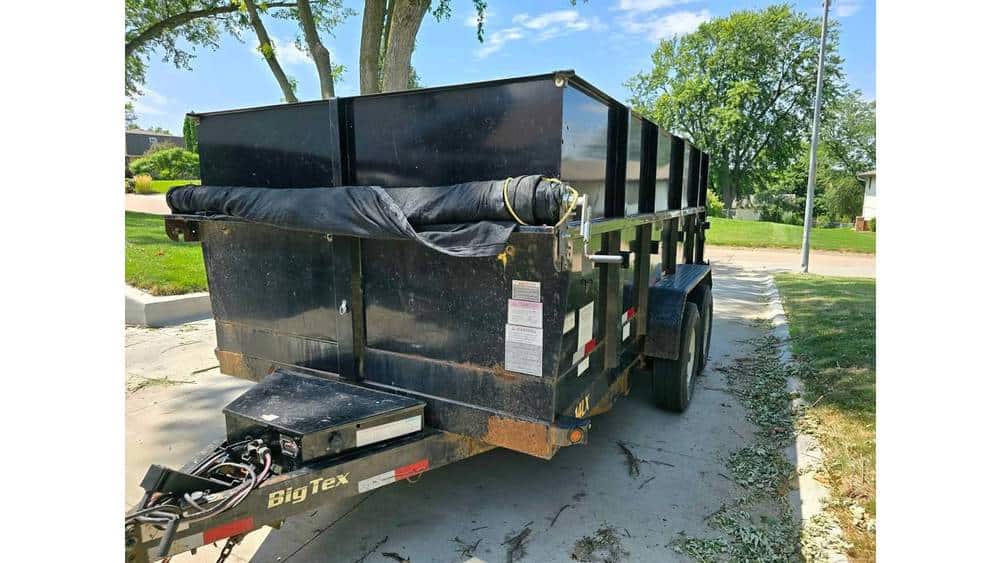 Heavy-duty black dump trailer with dual axles, parked on a paved surface in a green outdoor setting.