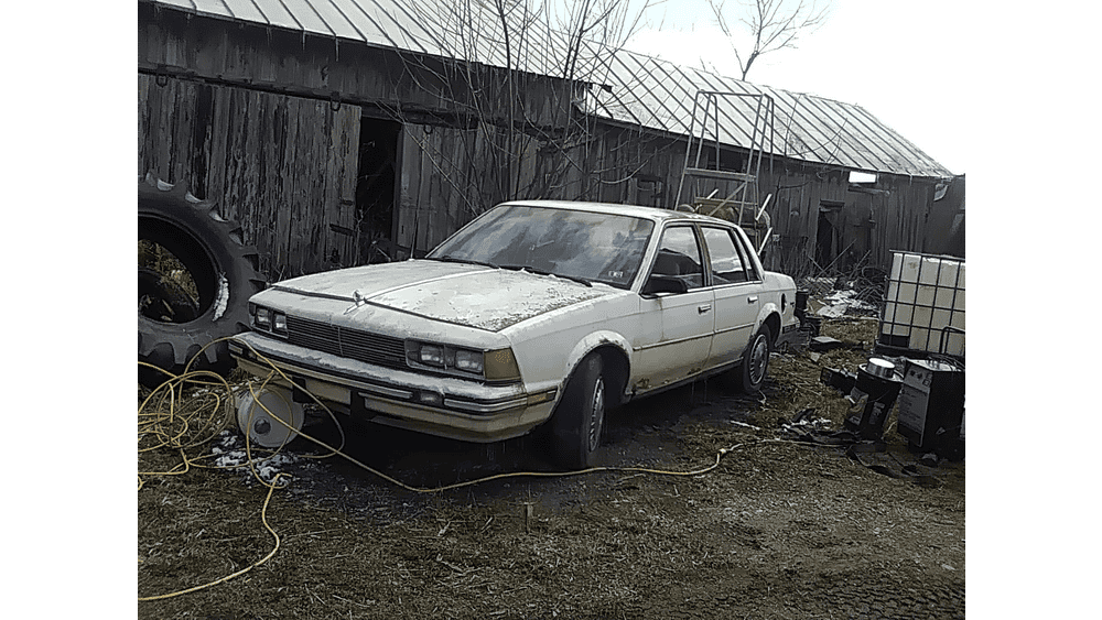 Old beige 1980s sedan parked in a rustic yard with wooden shed, tires, and equipment around.