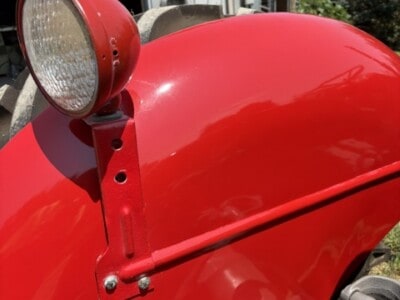 Red tractor with large front fender and round headlight, outdoors near a metal building.