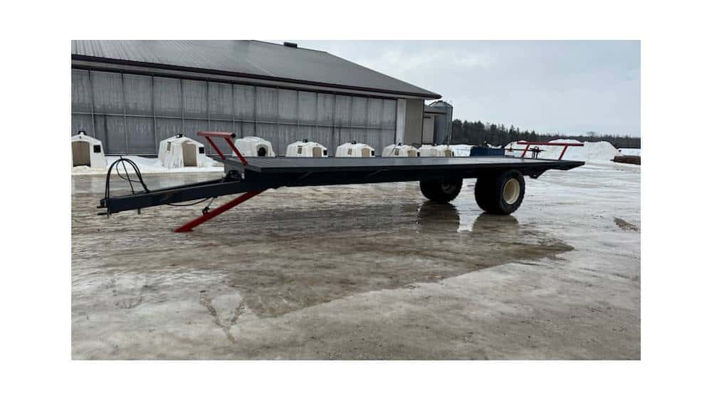 Flatbed trailer with dual axles parked outdoors on a snowy surface near a large barn.