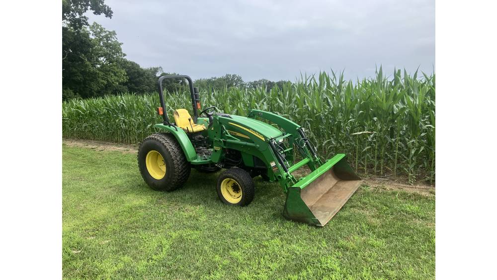 A.LT tag: Green John Deere tractor with front loader attachment on grassy field near cornfield, under cloudy sky.