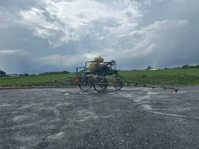 Farm sprayer on wheeled cart with tank and spray booms under cloudy sky.