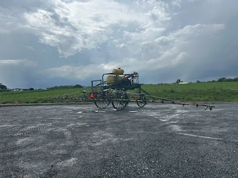 Farm sprayer on wheeled cart with tank and spray booms under cloudy sky.