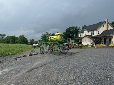 Large yellow and green agricultural sprayer with tank and booms on gravel driveway.