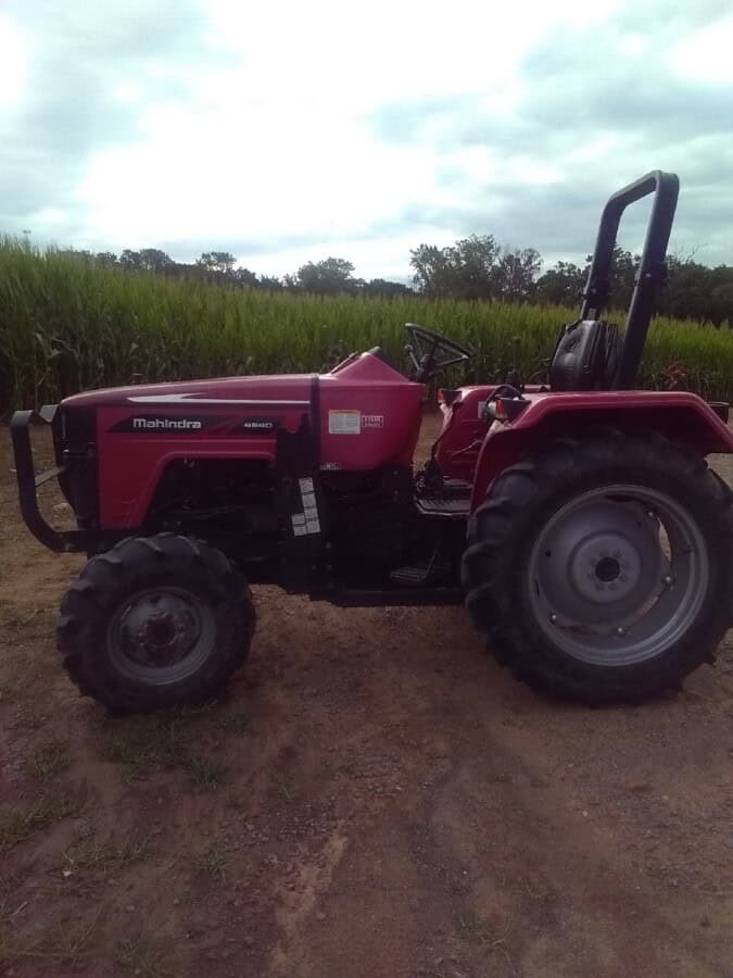 Mahindra tractor from 2020 model year, red color, parked on farm field with green crops and cloudy sky in background.
