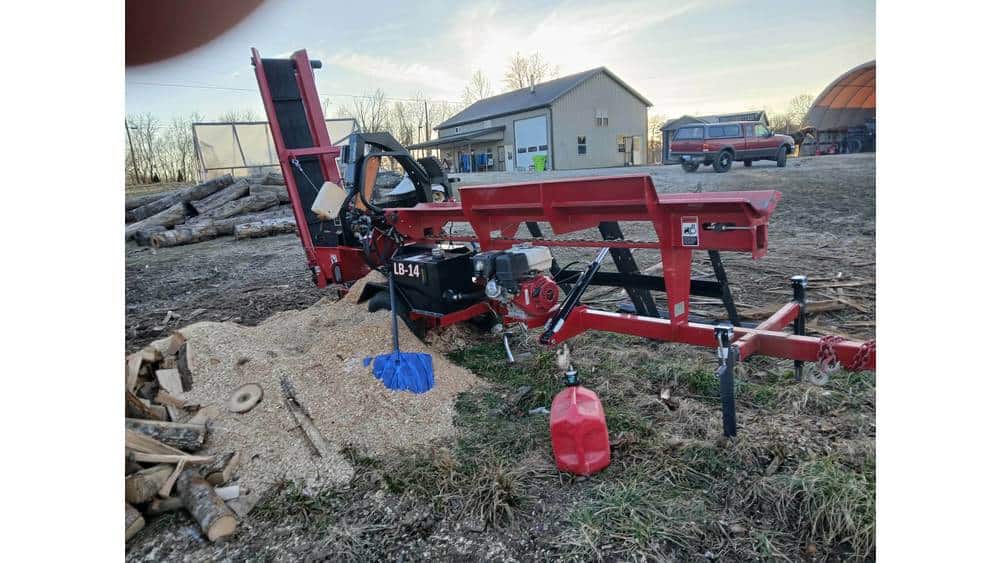 Red log splitter machine on a farm with a house and trees in the background during daytime.