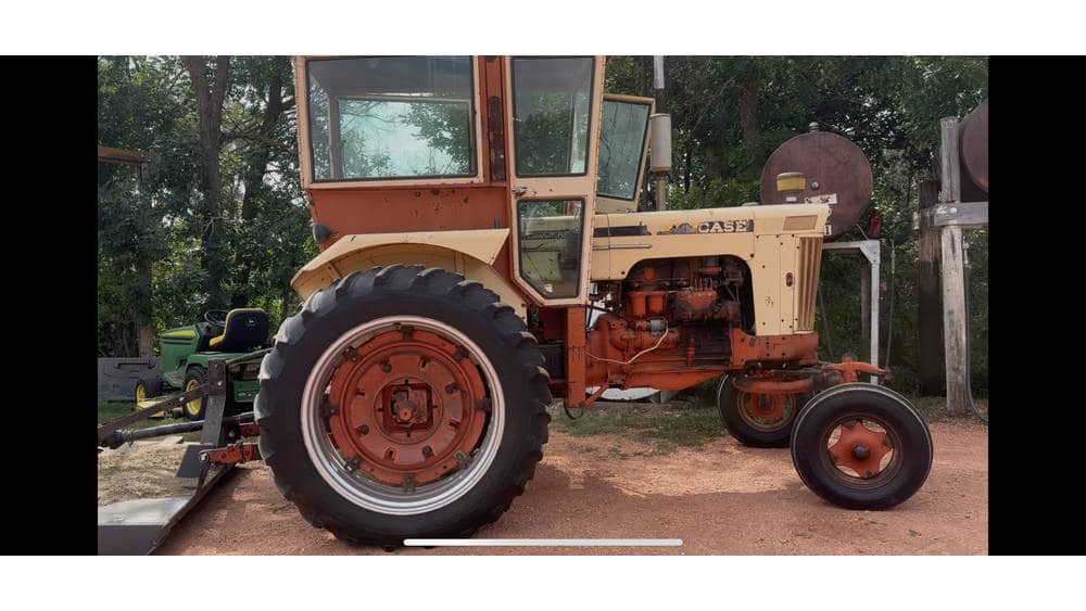 Vintage 1960s beige tractor with large rear tires and enclosed cab, parked outdoors among trees.