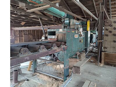 Industrial woodworking machine in a workshop setting, showing metal components and conveyor system.