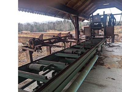 Industrial sawmill equipment with conveyor system under a wooden shelter, outdoors, no specific year or model visible.