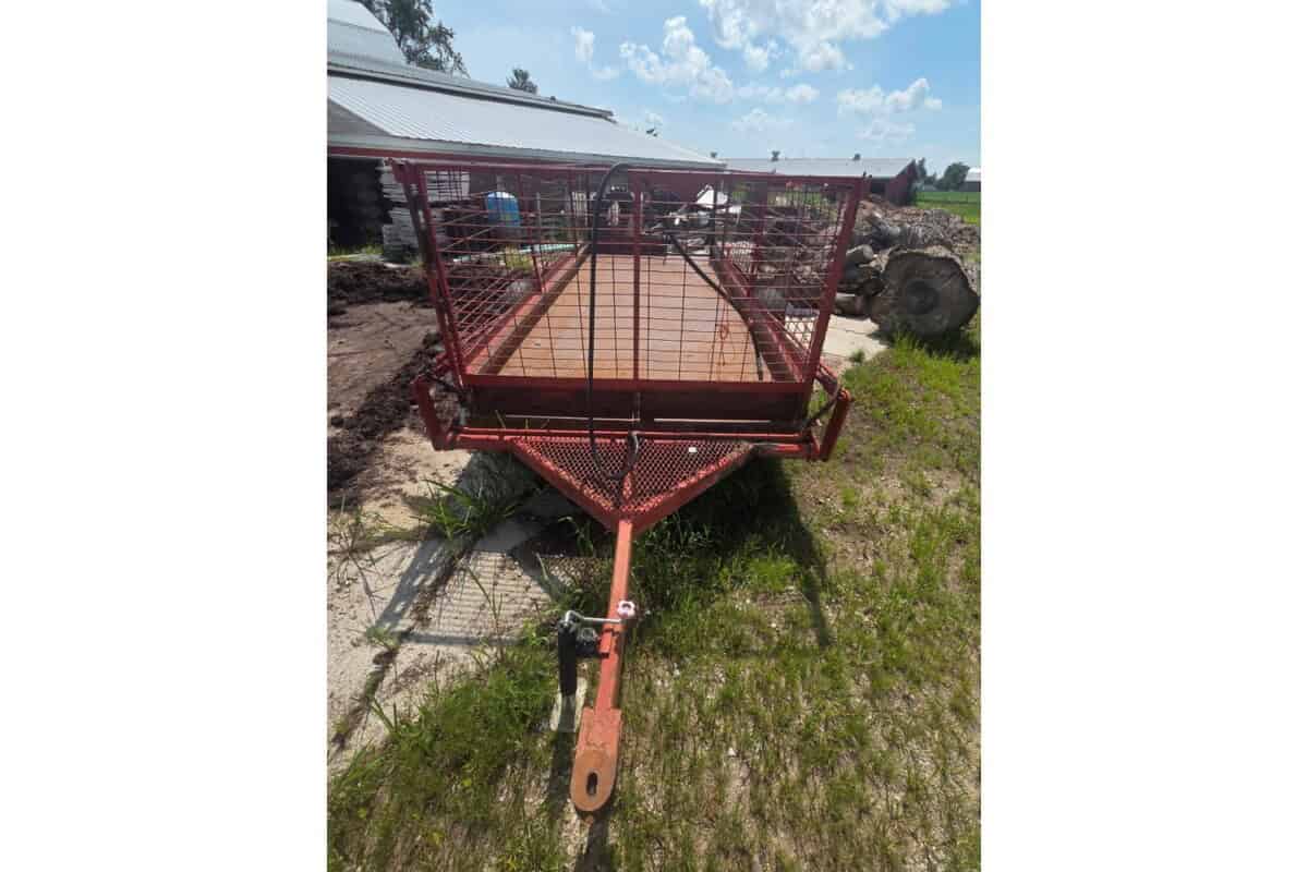 Red utility trailer with mesh sides and wooden floor, parked outdoors on grass.