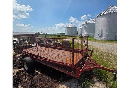 Heavy-duty red farm trailer parked on gravel with grain silos in the distance under a partly cloudy sky.