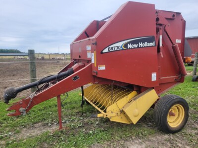 New Holland BR730 round baler for hay and straw collection, red color, with yellow pickup mechanism, on a farm field.