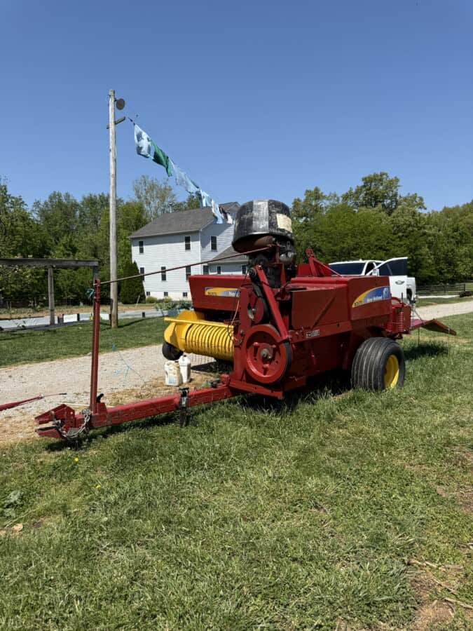 Red hay baler machine with yellow pickup and black engine, outdoors on green grass under clear blue sky.