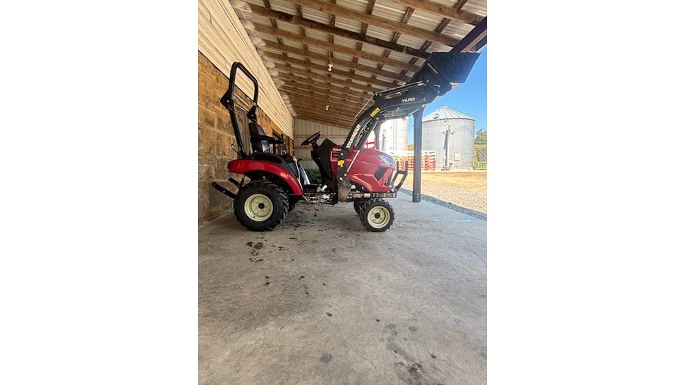 Red compact tractor with front loader attachment parked under a wooden shelter.