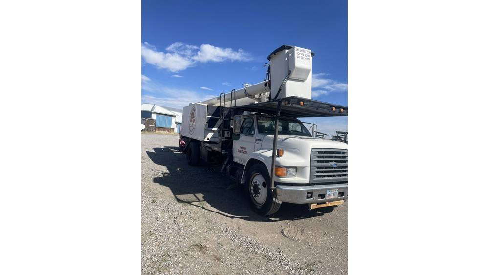 White bucket truck with aerial lift, likely from 2000s, parked outdoors on gravel under blue sky.