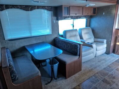 Interior view of a recreational vehicle with seating and dining area, featuring two armchairs, a booth-style dinette, and lar.