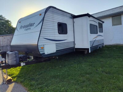 Prowler Lynx travel trailer, white with blue accents, parked on grass near a building, outdoor camping setup.