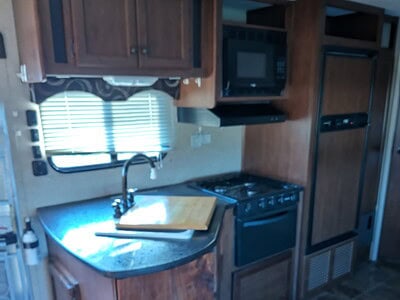 Compact kitchen area with wooden cabinets, stove, microwave, and window in a recreational vehicle.