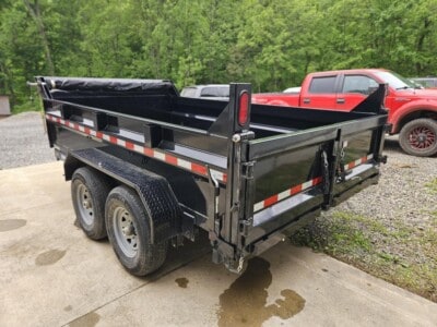 Black dump trailer with dual axles and red and white reflective tape, parked outdoors with trees in the background.