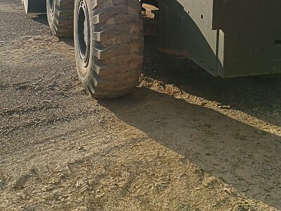 Excavator with a black cab and large tires parked outdoors on a dirt surface, surrounded by trees and a building in the backg.