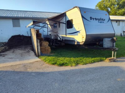Prowler Lynx travel trailer parked on grass near a building, with a pile of dirt and hay bales nearby.