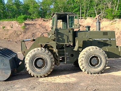 Excavator loader with a large bucket, green color, on a construction site with dirt and trees in the background.