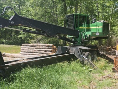Green forestry harvester with long arm and cutting head in a wooded area, used for logging operations.