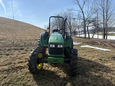 John Deere 5055E tractor on a farm field during winter, with leafless trees and snow patches in the background.