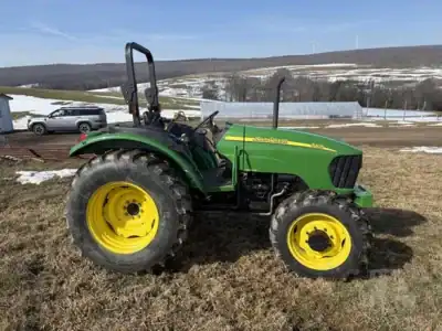John Deere 5075E tractor with green body and yellow wheels, parked on grassy field with snow-capped hills in background.