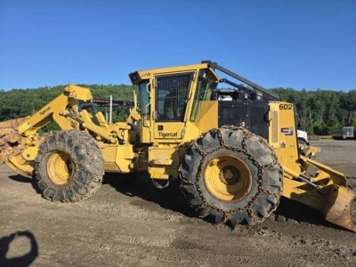 John Deere 602 Tigercat forestry skidder with large tires and cab, yellow and black color, on a dirt lot.
