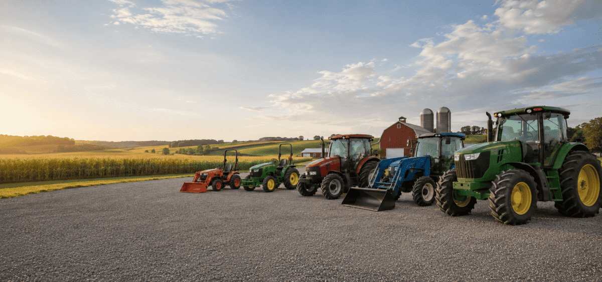 Multiple tractors and equipment parked on gravel with a rural landscape and sunset sky.