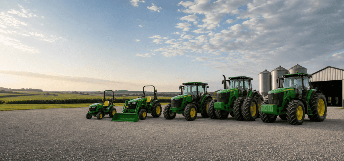 Lineup of green John Deere tractors and equipment on a farm with blue sky and clouds.