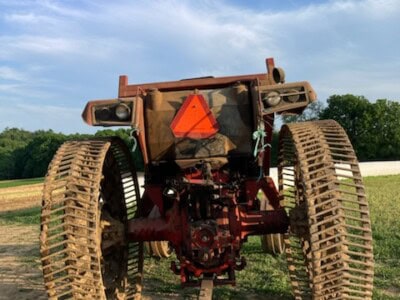 Old Caterpillar tracked tractor with rust and a reflective triangle on the back, outdoors on a dirt field under a blue sky.