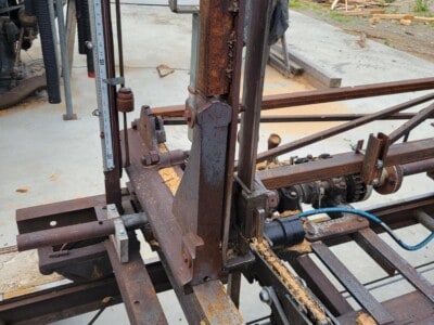 Rusty circular sawmill machine with chains and metal frame for lumber processing.
