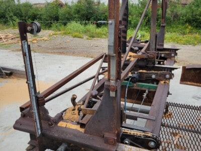 Rusty circular sawmill with metal frame and cutting blade, outdoor setting with storage buildings in background.