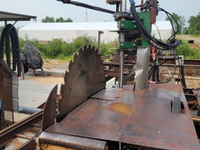 Rusty circular saw mill with hydraulic arm and control box on outdoor railway track.