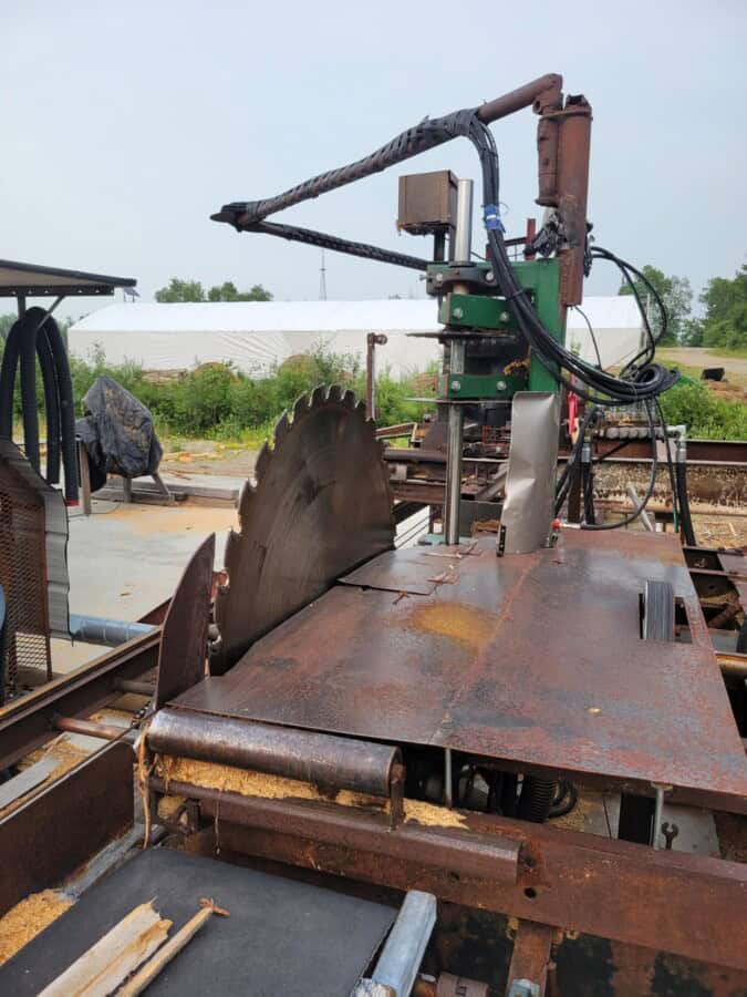 Rusty circular saw mill with hydraulic arm and control box on outdoor railway track.