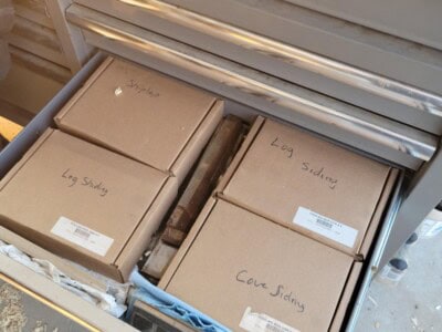 Four labeled cardboard storage boxes in a workshop drawer for logs and siding.
