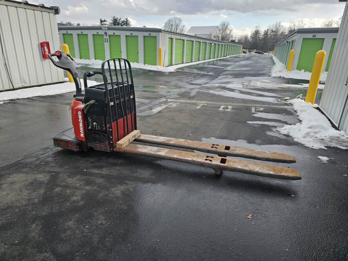 2006 Raymond 8400 electric pallet jack with black handle and red body on wet asphalt in storage facility.