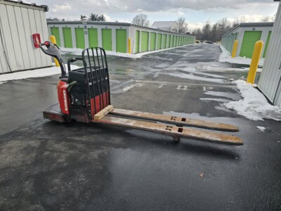 2006 Raymond 8400 electric pallet jack with black handle and red body on wet asphalt in storage facility.