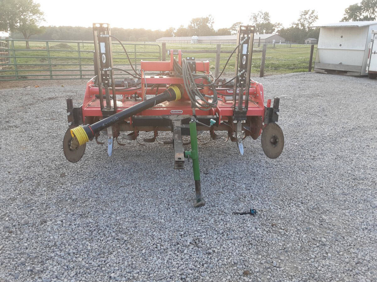 Red farm equipment with hydraulic hoses and wheels on gravel surface.
