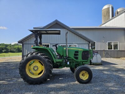 Green John Deere 6100D tractor with large rear tires and cab, parked outside a farm building on gravel surface.