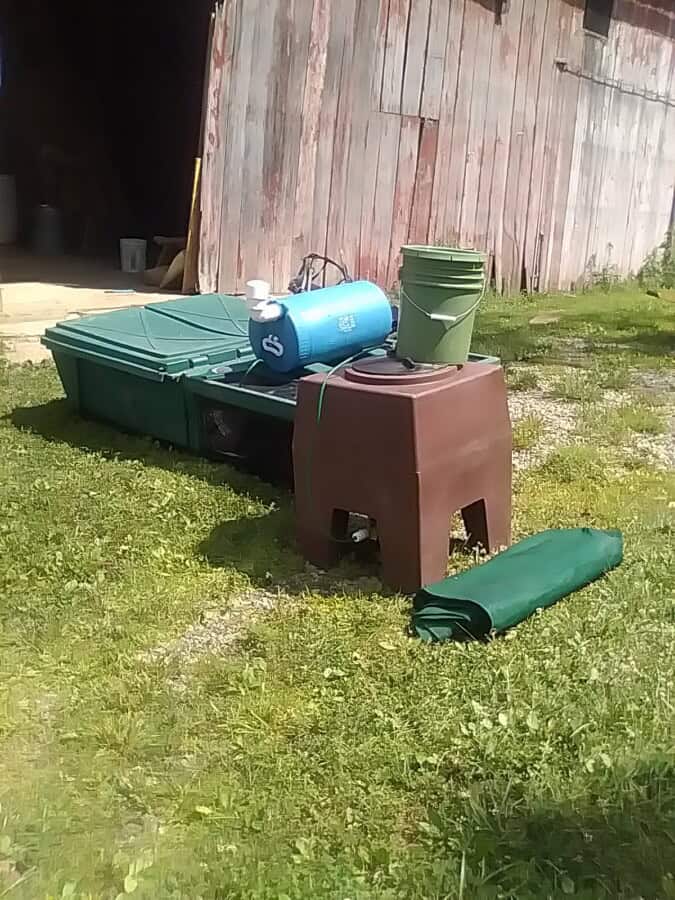 Assorted outdoor storage bins, buckets, and a rolled-up tarp on grass near a wooden shed.