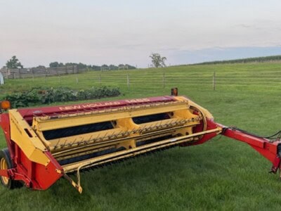 Red and yellow haybine with cutting blades on green field under blue sky.
