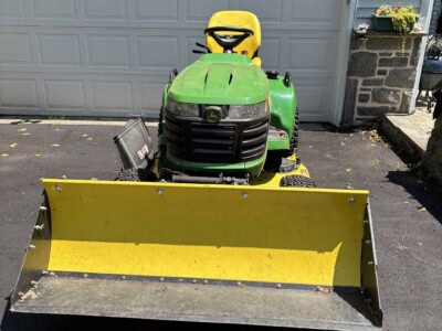 Green John Deere lawn tractor with yellow snow plow attachment in driveway.