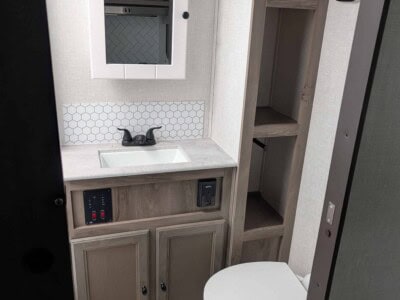 Small RV bathroom with sink, mirror, toilet, and storage shelves in neutral tones.