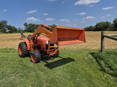 Orange compact utility loader with bucket attachment on grassy field under blue sky.