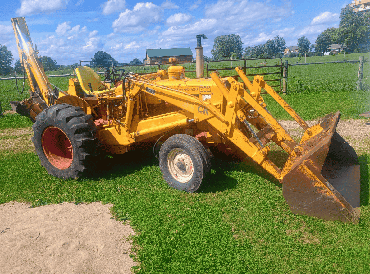 Yellow Case 580 backhoe loader with front bucket and rear excavator arm on grassy field.